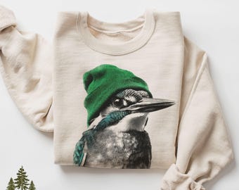 May include: A sand-coloured sweatshirt with a detailed illustration of a bird wearing a green beanie. The bird has blue and white markings. The sweatshirt has long sleeves and a crew neck. The word "Sand" is in the upper left corner.
