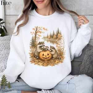 May include: A white crewneck sweatshirt featuring a graphic print of three raccoons gathered around a carved jack-o'-lantern pumpkin in an autumn forest setting with golden trees, fallen leaves, and a river, with the text Old Pines Co. at the bottom.