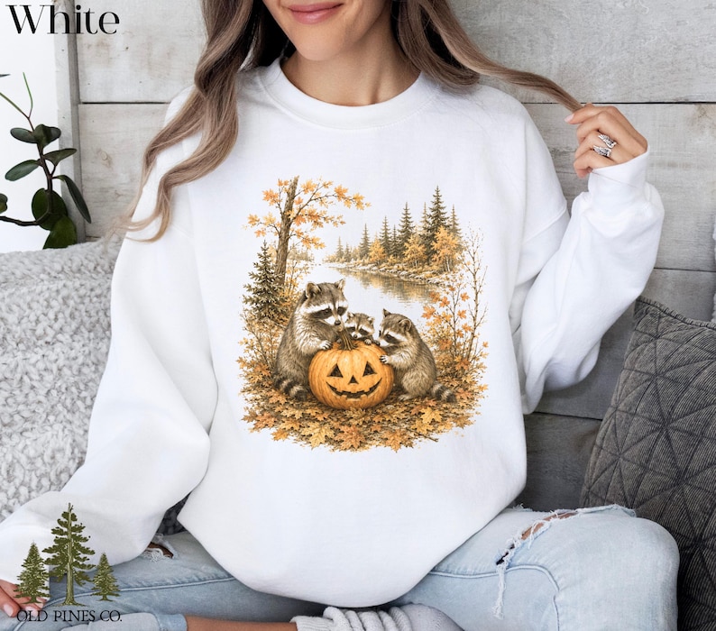 May include: A white crewneck sweatshirt featuring a graphic print of three raccoons gathered around a carved jack-o'-lantern pumpkin in an autumn forest setting with golden trees, fallen leaves, and a river, with the text Old Pines Co. at the bottom.