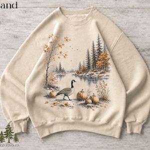 Puede incluir: Una sudadera de cuello redondo color arena con un gráfico impreso de un ganso canadiense junto a un lago rodeado de pinos, follaje otoñal y varias calabazas. El texto "Sand" aparece en la esquina superior izquierda de la imagen.