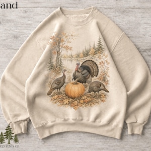 May include: A sand-colored crewneck sweatshirt featuring a detailed graphic print of three turkeys standing around a large orange pumpkin in a forest setting with autumn leaves, trees, and a lake in the background. The word Sand is printed above.