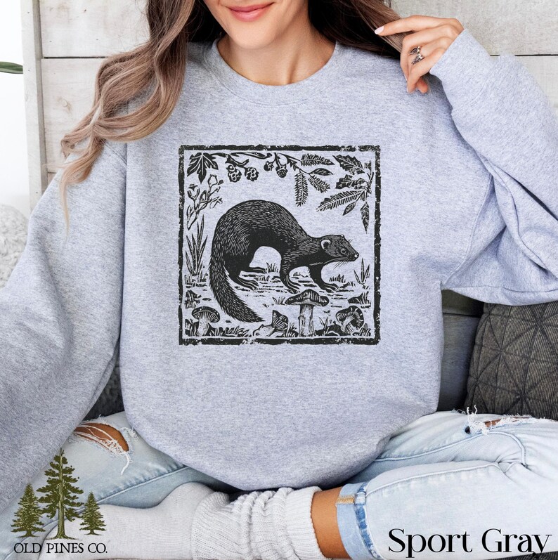 May include: Light gray sweatshirt with a black and white illustration of a ferret in a natural setting. The design is framed with botanical elements. The words "OLD PINES CO." are printed at the bottom left, and "Sport Gray" is printed at the bottom right.