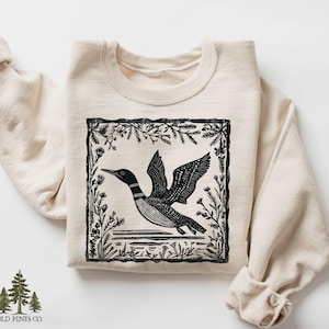 May include: Cream-colored sweatshirt featuring a black and white illustration of a loon in flight, framed by floral and plant elements. The sweatshirt has a crew neck and long sleeves. The brand logo, "OLD PINES CO." is at the bottom left.