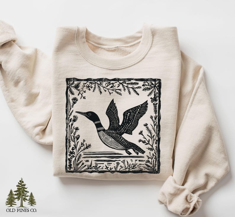 May include: Cream-colored sweatshirt featuring a black and white illustration of a loon in flight, framed by floral and plant elements. The sweatshirt has a crew neck and long sleeves. The brand logo, "OLD PINES CO." is at the bottom left.