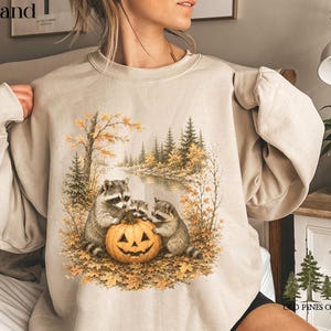 May include: A sand-colored crewneck sweatshirt featuring a graphic print of three raccoons gathered around a carved jack-o'-lantern pumpkin. The scene is set in an autumn forest with a river, pine trees, and fallen leaves in shades of orange and brown.