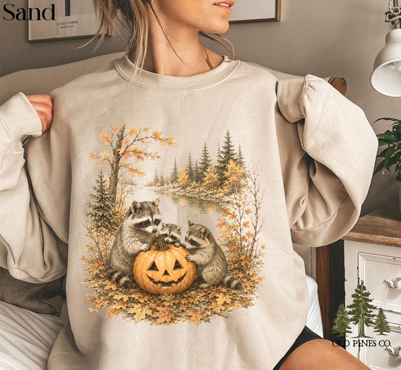 May include: A sand-colored crewneck sweatshirt featuring a graphic print of three raccoons gathered around a carved jack-o'-lantern pumpkin. The scene is set in an autumn forest with a river, pine trees, and fallen leaves in shades of orange and brown.