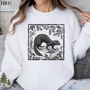 May include: White sweatshirt featuring a black and white illustration of a ferret surrounded by plants and mushrooms within a rectangular frame. The word "White" is printed at the top left. The brand logo "OLD PINES CO." is at the bottom left.