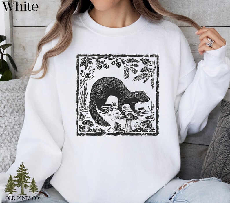 May include: White sweatshirt featuring a black and white illustration of a ferret surrounded by plants and mushrooms within a rectangular frame. The word "White" is printed at the top left. The brand logo "OLD PINES CO." is at the bottom left.