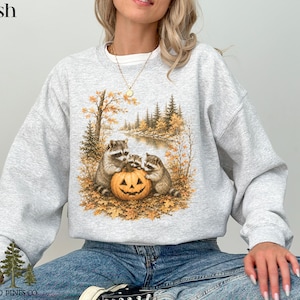 May include: An ash gray crewneck sweatshirt featuring a graphic print of three raccoons gathered around a carved jack-o'-lantern pumpkin. The scene is set in an autumn forest with golden leaves, pine trees, and a river in the background.