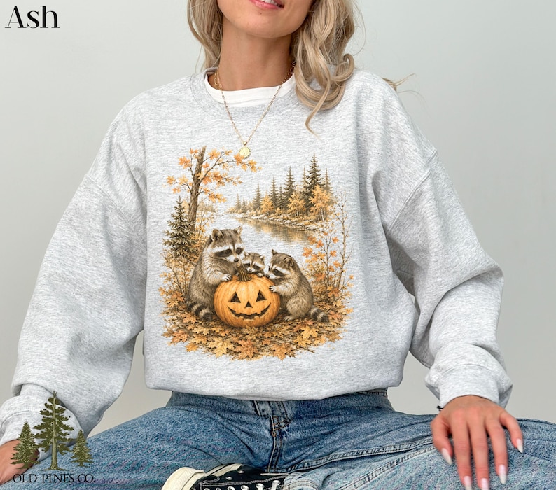 May include: An ash gray crewneck sweatshirt featuring a graphic print of three raccoons gathered around a carved jack-o'-lantern pumpkin. The scene is set in an autumn forest with golden leaves, pine trees, and a river in the background.