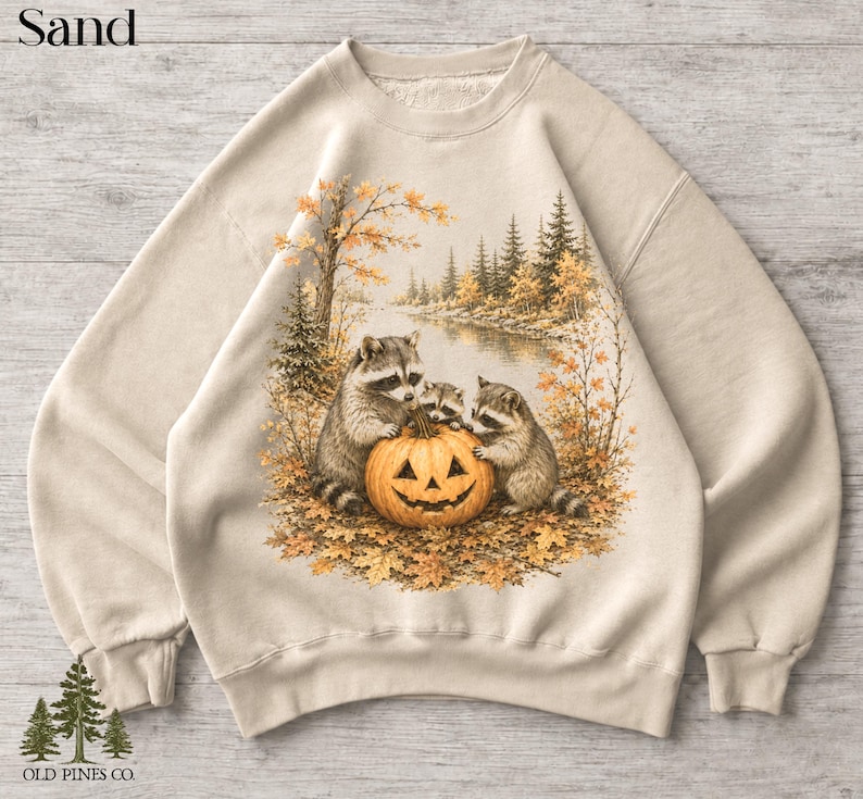 May include: A sand-colored crewneck sweatshirt featuring a graphic print of three raccoons gathered around a carved jack-o'-lantern pumpkin. The scene is set in an autumn forest with golden leaves, pine trees, and a river. The text Sand is visible.