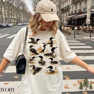 May include: Ivory-colored t-shirt featuring a detailed illustration of loons in a water scene. The shirt has the word "IVORY" printed at the bottom left and the "OLD PINES CO." logo at the bottom right. A beige baseball cap with a black logo is also visible.