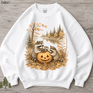 May include: A white crewneck sweatshirt featuring a graphic print of three raccoons gathered around a carved jack-o'-lantern pumpkin. The scene includes a forest landscape with autumn foliage, trees, and a river. The Gildan 18000 label is visible.