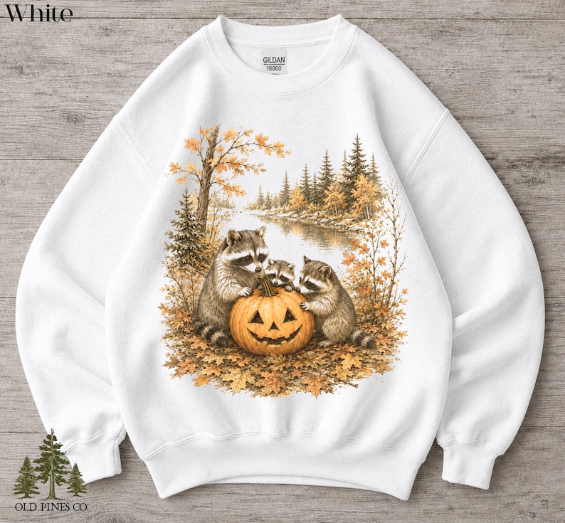 May include: A white crewneck sweatshirt featuring a graphic print of three raccoons gathered around a carved jack-o'-lantern pumpkin. The scene includes a forest landscape with autumn foliage, trees, and a river. The Gildan 18000 label is visible.
