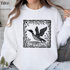 May include: White sweatshirt featuring a black and white illustration of a loon in flight, framed by a botanical border. The sweatshirt has the text "White" at the top and the "Old Pines Co." logo at the bottom.