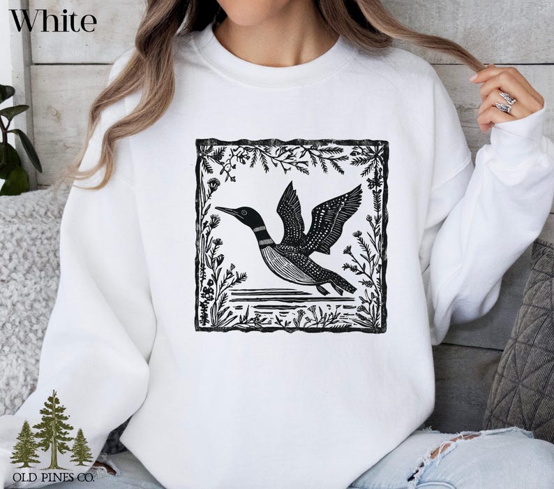 May include: White sweatshirt featuring a black and white illustration of a loon in flight, framed by a botanical border. The sweatshirt has the text "White" at the top and the "Old Pines Co." logo at the bottom.