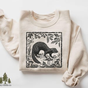 May include: A cream-colored sweatshirt featuring a black and white illustration of a ferret in a forest scene with mushrooms and foliage. The design is framed within a black border. The brand logo "OLD PINES CO." is at the bottom.