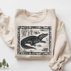 May include: A cream-colored sweatshirt featuring a black and white illustration of an alligator in a swamp scene. The design includes plants and lily pads within a decorative border. The sweatshirt has a crew neck and long sleeves.