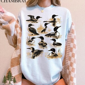 May include: Light blue sweatshirt featuring a detailed illustration of various loons in a water scene. The design includes multiple loon depictions in different poses, with the word "CHAMBRAY" at the top and "OLD PINES CO." at the bottom.