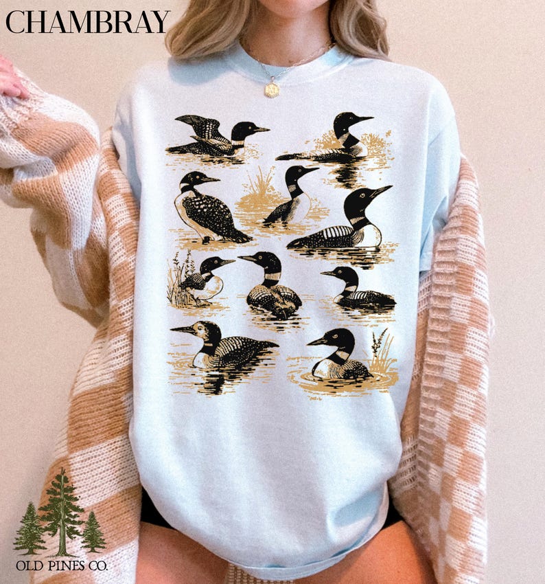 May include: Light blue sweatshirt featuring a detailed illustration of various loons in a water scene. The design includes multiple loon depictions in different poses, with the word "CHAMBRAY" at the top and "OLD PINES CO." at the bottom.