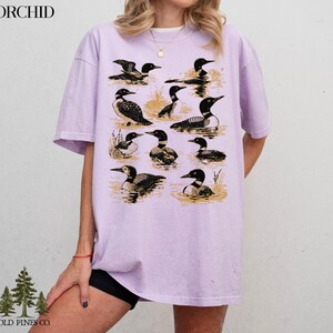 May include: A light purple t-shirt featuring a graphic of various loons in a water scene. The loons are depicted in black and white, with a vintage aesthetic. The shirt has the word "ORCHID" printed at the top left and "OLD PINES CO." at the bottom left.