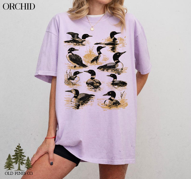 May include: A light purple t-shirt featuring a graphic of various loons in a water scene. The loons are depicted in black and white, with a vintage aesthetic. The shirt has the word "ORCHID" printed at the top left and "OLD PINES CO." at the bottom left.
