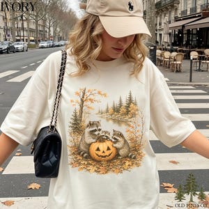 May include: An ivory t-shirt featuring a graphic print of three raccoons gathered around a carved jack-o'-lantern pumpkin in an autumn forest setting with fall foliage, pine trees, and a river, paired with a black quilted shoulder bag.