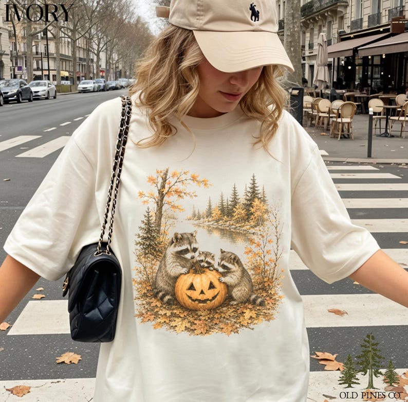May include: An ivory t-shirt featuring a graphic print of three raccoons gathered around a carved jack-o'-lantern pumpkin in an autumn forest setting with fall foliage, pine trees, and a river, paired with a black quilted shoulder bag.