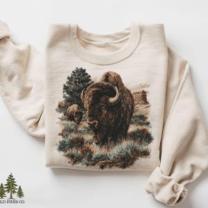 May include: A cream-colored sweatshirt featuring a detailed illustration of two bison in a grassy landscape with a tree and rock formations. The graphic is in shades of brown, green, and tan. The brand name "OLD PINES CO." is printed at the bottom.