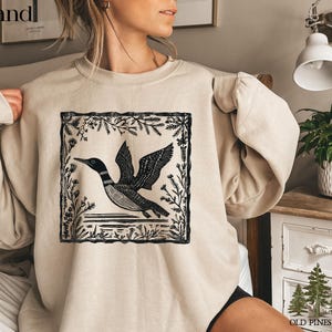 May include: A sand-colored sweatshirt featuring a black and white graphic of a loon in flight, framed by botanical elements. The design is centered on the front of the long-sleeved top. The text "Sand" is in the upper left corner.