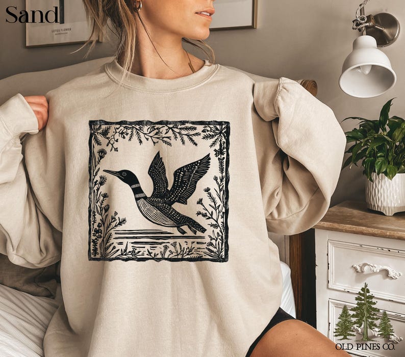 May include: A sand-colored sweatshirt featuring a black and white graphic of a loon in flight, framed by botanical elements. The design is centered on the front of the long-sleeved top. The text "Sand" is in the upper left corner.
