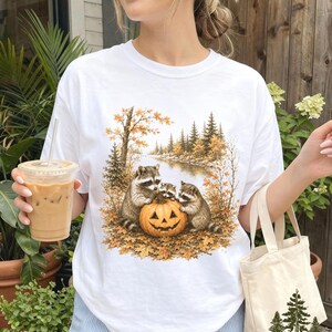 May include: A white t-shirt featuring a graphic print of three raccoons gathered around a carved jack-o'-lantern in an autumn forest setting with golden leaves, pine trees, and a river, plus a small canvas tote bag with a pine tree logo.