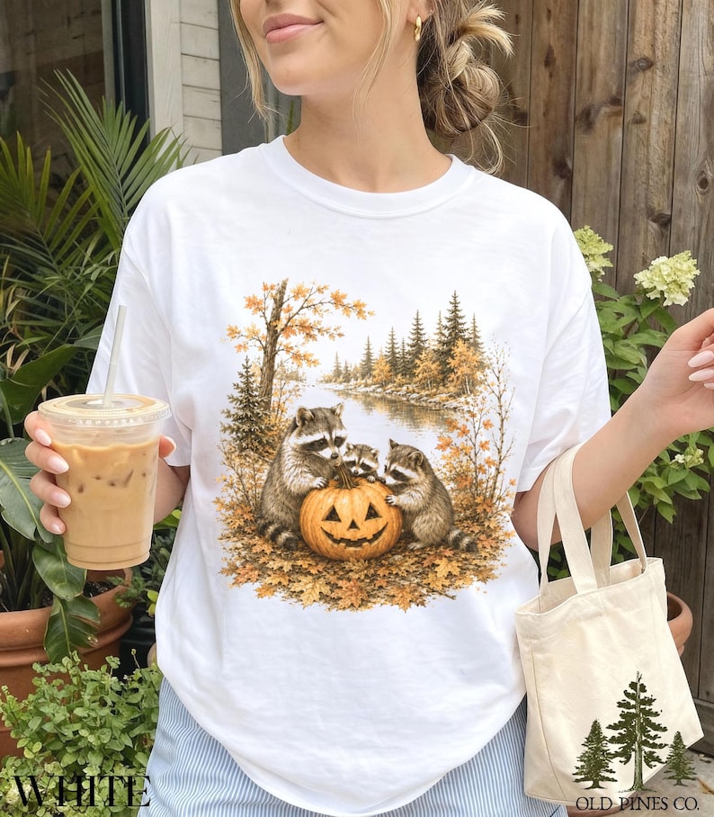May include: A white t-shirt featuring a graphic print of three raccoons gathered around a carved jack-o'-lantern in an autumn forest setting with golden leaves, pine trees, and a river, plus a small canvas tote bag with a pine tree logo.