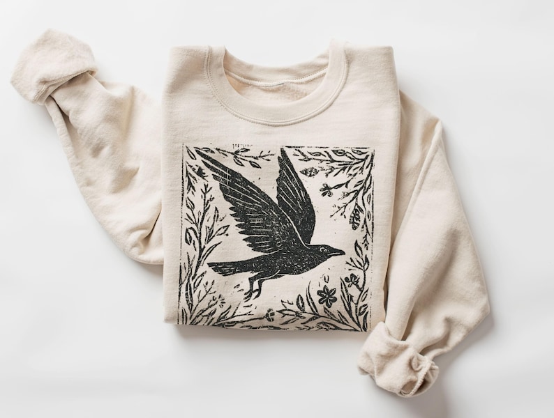 May include: A cream-colored sweatshirt with a black graphic of a bird in flight, framed by botanical elements. The bird illustration is centered on the front of the folded sweatshirt. The sleeves are rolled up.
