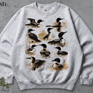May include: Ash-colored sweatshirt featuring a detailed illustration of multiple loons in various poses. The design includes loons swimming and standing in water, with a vintage aesthetic. The text "OLD PINES CO." is at the bottom.