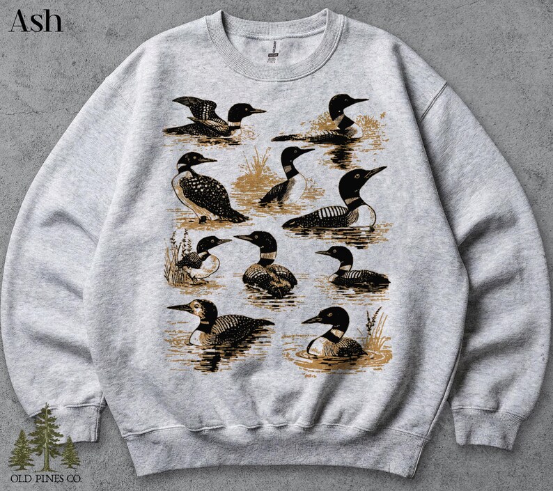 May include: Ash-colored sweatshirt featuring a detailed illustration of multiple loons in various poses. The design includes loons swimming and standing in water, with a vintage aesthetic. The text "OLD PINES CO." is at the bottom.