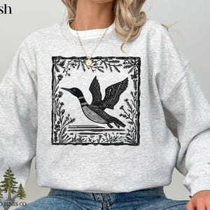 May include: Ash-colored sweatshirt featuring a black and white graphic of a loon in flight, framed by floral elements. The sweatshirt has a crew neck and long sleeves. The text "OLD PINES CO." is printed at the bottom left.