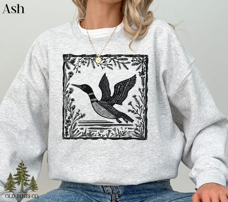 May include: Ash-colored sweatshirt featuring a black and white graphic of a loon in flight, framed by floral elements. The sweatshirt has a crew neck and long sleeves. The text "OLD PINES CO." is printed at the bottom left.