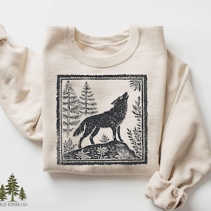 May include: A cream-colored sweatshirt featuring a black and white graphic of a howling wolf in a forest scene. The design is framed within a rectangular border. The brand name "OLD PINES CO." is printed at the bottom left.