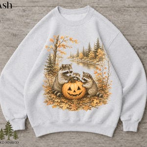 May include: An ash gray crewneck sweatshirt featuring a graphic print of three raccoons gathered around a carved jack-o'-lantern pumpkin. The scene includes autumn foliage, trees, and a river. The word Ash is printed in the top left corner.
