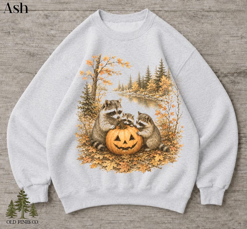 May include: An ash gray crewneck sweatshirt featuring a graphic print of three raccoons gathered around a carved jack-o'-lantern pumpkin. The scene includes autumn foliage, trees, and a river. The word Ash is printed in the top left corner.