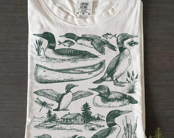 Könnte beinhalten: Weißes T-Shirt mit einer grünen Illustration von Eistauchern, Fischen, einem Kanu und einer Hütte am See. Das Shirt hat einen Rundhalsausschnitt und der Markenname "Comfort Colors" ist auf dem Etikett sichtbar. Das Shirt ist sauber gefaltet.