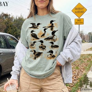 May include: Sage green t-shirt featuring a detailed illustration of loons in various poses. The design includes black and white loons with orange accents, set against a water-like background. The shirt has the text "BAY" at the top left.