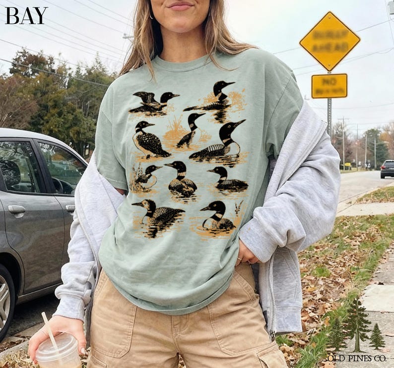 May include: Sage green t-shirt featuring a detailed illustration of loons in various poses. The design includes black and white loons with orange accents, set against a water-like background. The shirt has the text "BAY" at the top left.