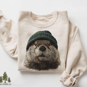 May include: Cream-coloured sweatshirt with a detailed illustration of a groundhog wearing a dark green beanie. The groundhog's face is centered on the front of the folded sweatshirt. The sweatshirt has long sleeves and a crew neck.
