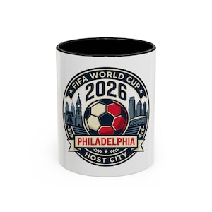 May include: A white ceramic mug with a black interior and rim, featuring a circular graphic for the 2026 FIFA World Cup in Philadelphia. The design includes a soccer ball, city skyline silhouettes, and the text FIFA WORLD CUP 2026 PHILADELPHIA.