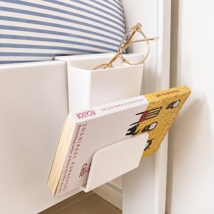 Könnte beinhalten: Ein weißer Nachttisch-Organizer mit eingebautem Buchhalter und einem kleinen Fach für Brillen. Ein Buch mit dem Titel "Cafe" von Toshikazu Kawaguchi befindet sich im Halter. Goldgerahmte Brillen liegen im Fach. Ein gestreiftes Kissen ist im Hintergrund.