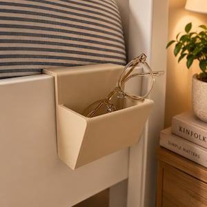 Op de afbeelding: Een beige nachtkastorganizer bevestigd aan een wit bedframe, met een bril erin. De organizer heeft een rechthoekige vorm met een klein compartiment. Een gestreept kussen is zichtbaar op de achtergrond.