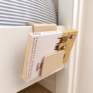 Puede incluir: Un sujetalibros de mesita de noche beige sujeto a un marco de cama blanco. Un libro titulado "the Cafe" de Toshikazu Kawaguchi se sostiene en el soporte. La cama tiene una almohada a rayas azules y blancas.