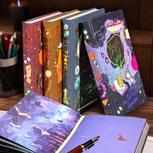 Könnte beinhalten: Eine Sammlung von Notizbüchern mit Fantasy-Thema und farbenfrohen Einbänden. Die Notizbücher zeigen Illustrationen von magischen Elementen, darunter ein Hexenhut, eine Schüssel mit Essen und die Worte "Magic World" und "The Fantasy Adventure".